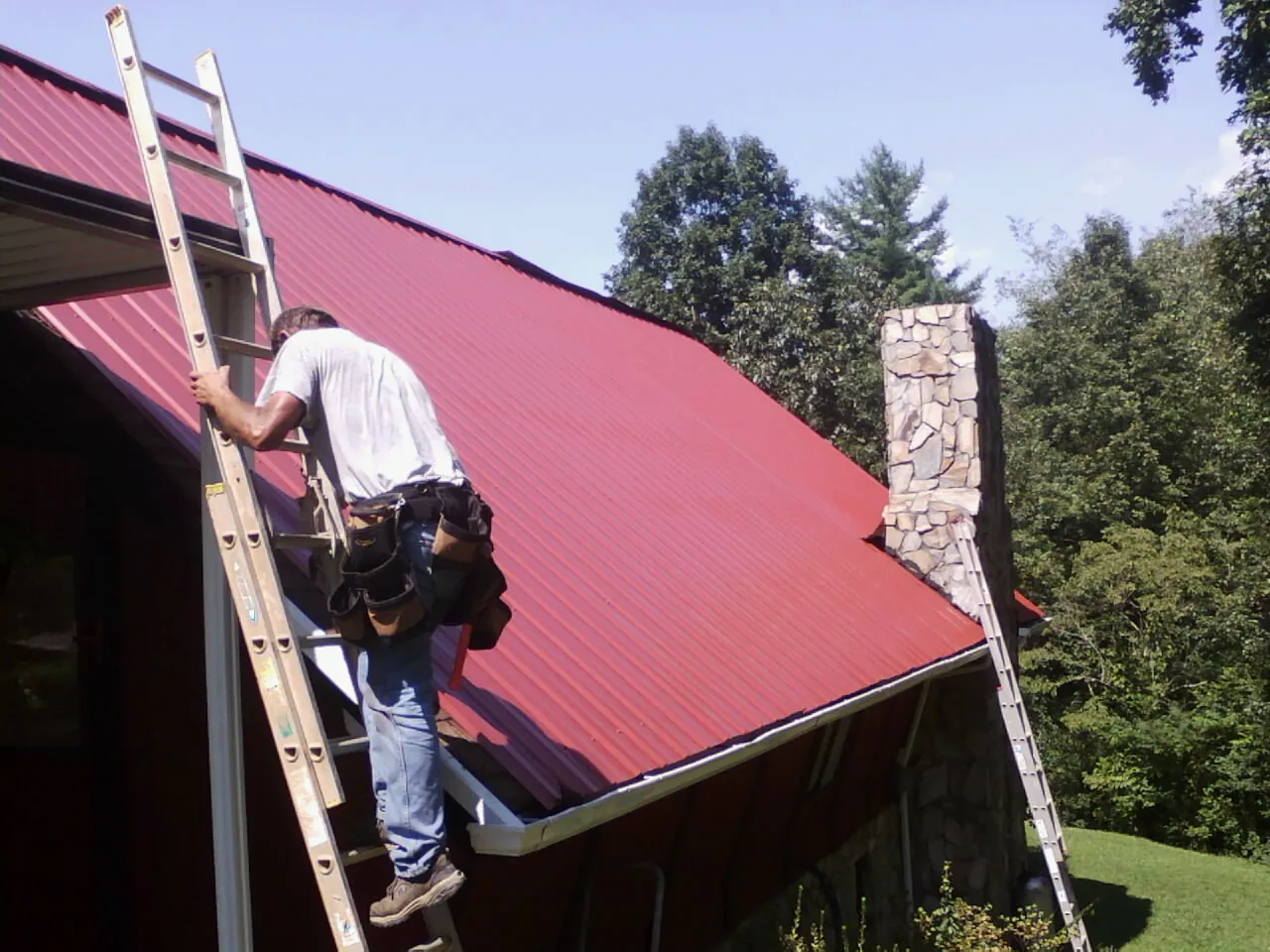 Roofer on a ladder beside a red metal roof and stone chimney — Soffit & Fascia Repair by Roofing Brothers in Airway Heights